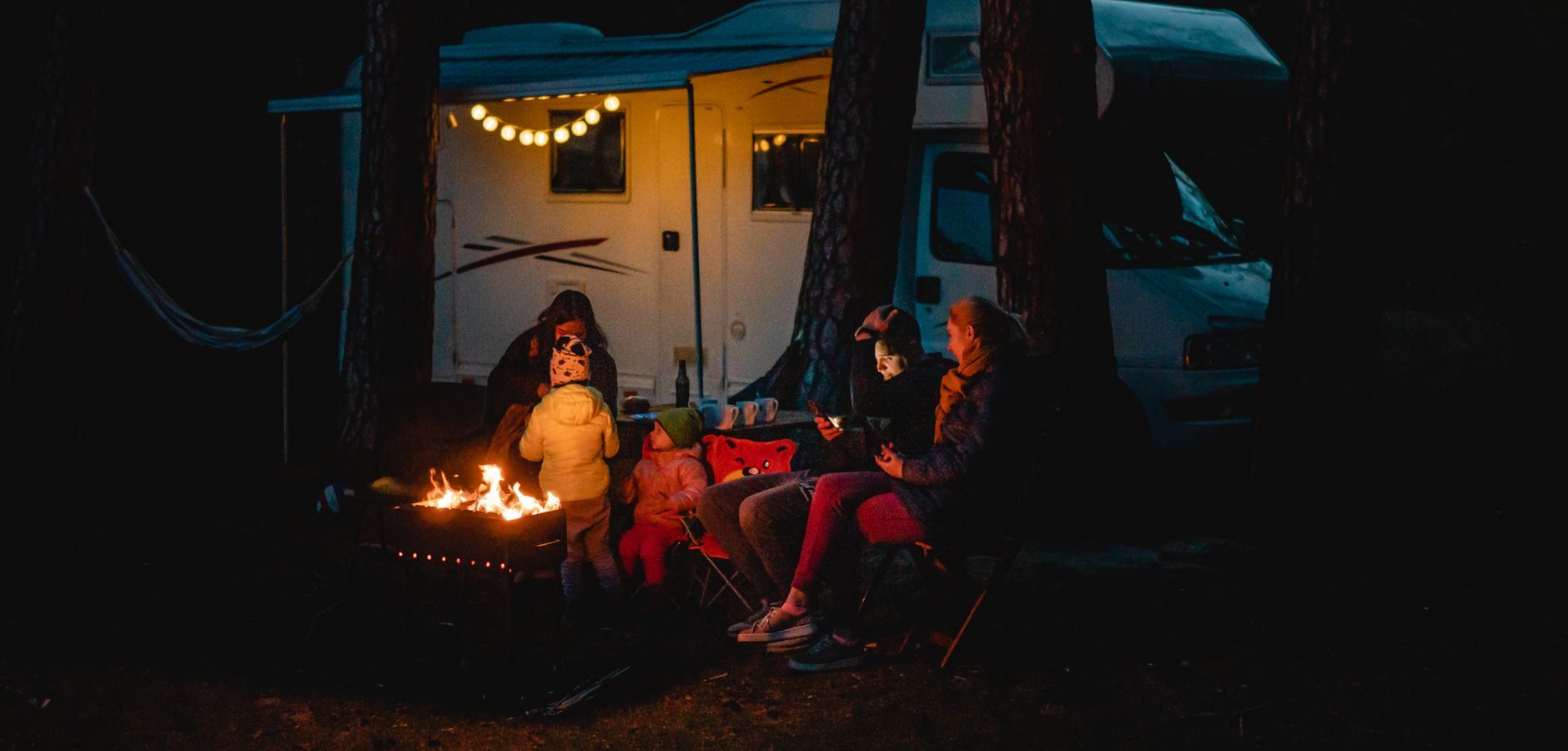 Camping-car blanc garé en forêt la nuit, une famille autour d’un feu de camp sous une guirlande lumineuse